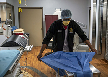Military personnel, wearing a mask, is spreading a sheet on an operating room bed