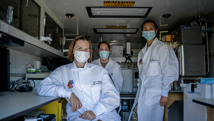 Image of Three military personnel, wearing masks and lab coats, pose for a picture in an Albanian lab. Click to open a larger version of the image.