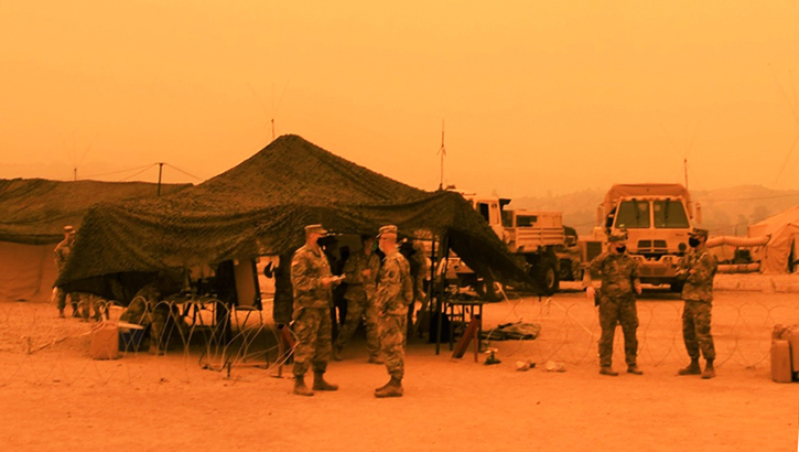 Image of Picture of a military tent; an orange, smoky hue surrounds the tent and soldiers. Click to open a larger version of the image.