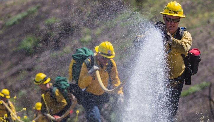 Image of Men in protective suits dousing a flame with water from a hose.