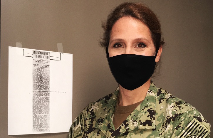 Image of Navy captain, wearing a mask, standing next to a piece of paper on the wall.