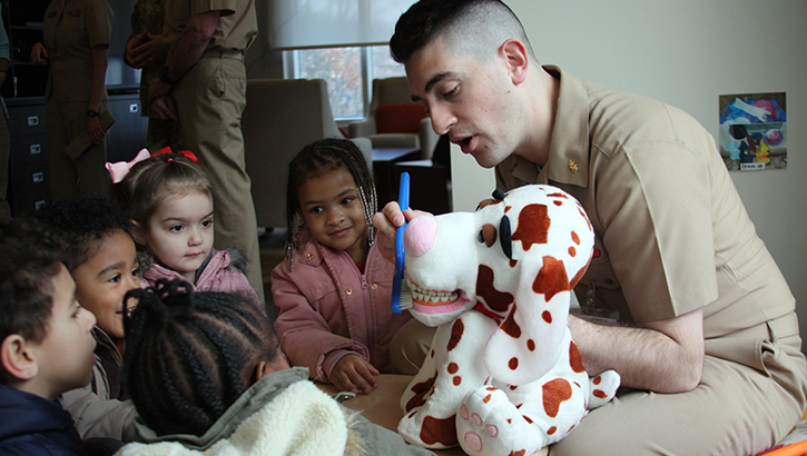Image of Military health personnel, sitting in front of a group of children, showing them how to brush their teeth using a stuffed animal. Click to open a larger version of the image.