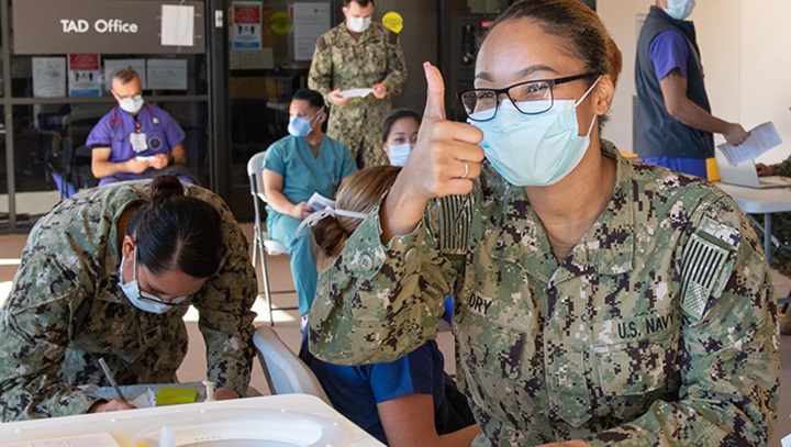 Image of Several military personnel, wearing masks, filling out paperwork. One woman is giving the thumbs up sign.