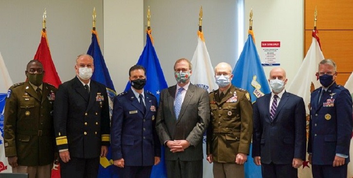Military personnel, wearing masks, standing in a line in front of flags