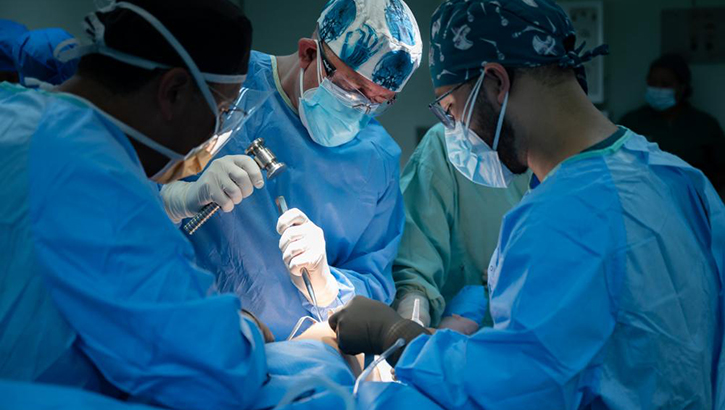 Image of Four surgeons in gowns, caps, and masks perform knee surgery.