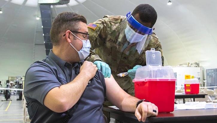 Medical director at Fort Riley, Kansas receives a COVID-19 vaccination In his left arm from a tech in personal protective equipment.