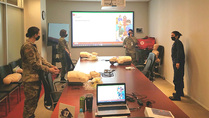 Image of Four military personnel in masks, in a conference room, looking at a presentation on a screen, with several practice dummies. Click to open a larger version of the image.