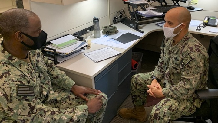 Image of Two military personnel, wearing masks, sitting at a desk talking.