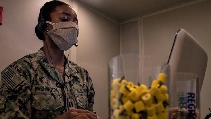 Image of Soldier wearing mask, sitting at laptop with a container of ear plugs close by.