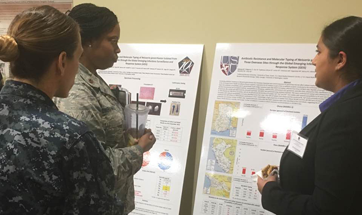 Nazia Rahman, right, manager of the Uniformed Services University’s Neisseria gonorrhoeae (GC) Repository, discusses a poster on the sexually-transmitted infection with U.S. Air Force Major Trinette Flowers-Torres, lead for GEIS’s Antimicrobial Resistance (AMR) Focus Area, left, and Navy Commander Franca Jones, front, chief of GEIS, examine poster on AMR surveillance. (AFHSB photo by Briana Booker).