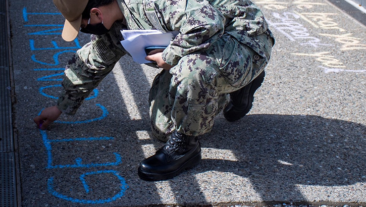 Image of Military personnel wearing a face mask writing messages with chalk on the ground. Click to open a larger version of the image.