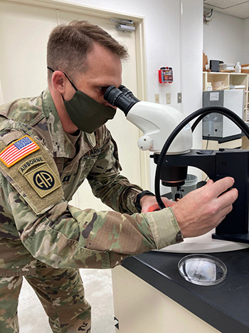 Military personnel wearing a face mask looking into a microscope