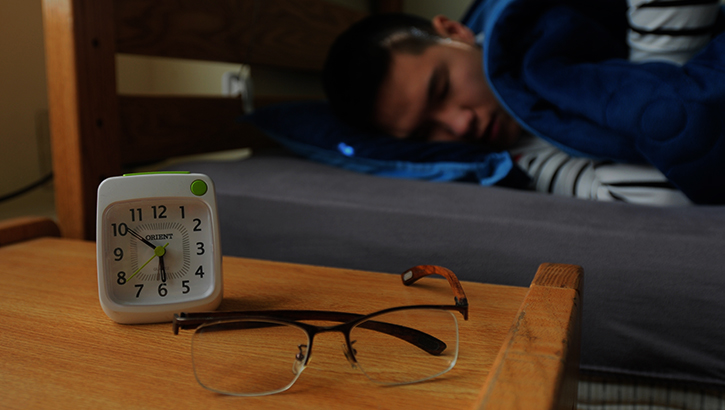 Image of a man sleeping in bed with alarm clock on side table. Click to open a larger version of the image. Click to open a larger version of the image.