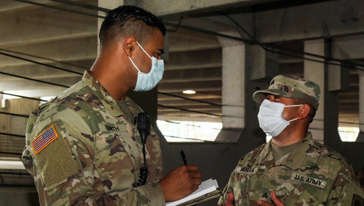 Two military personnel with mask on talking, while one is writing on a notepad