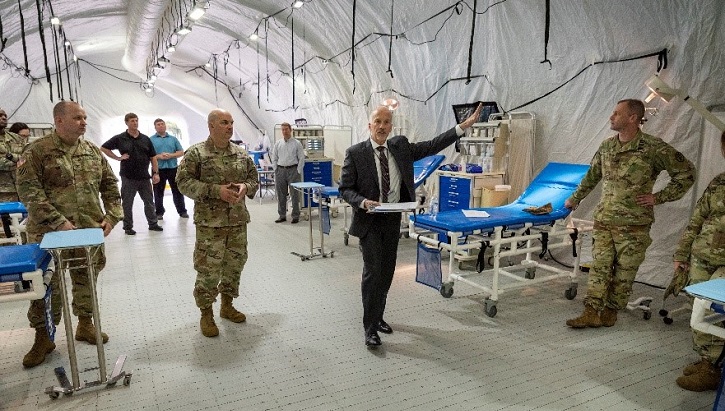 Image of soldiers and businessman in suit walking through an emergency shelter lined with beds and medical equipment