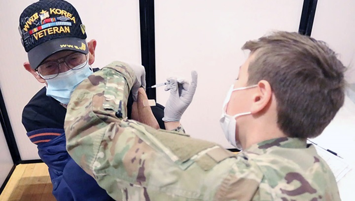 Image of Military personnel wearing a mask, giving the COVID-19 vaccine to a veteran wearing a mask.