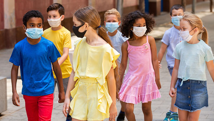 A bunch of children wearing face masks walk on a city street.