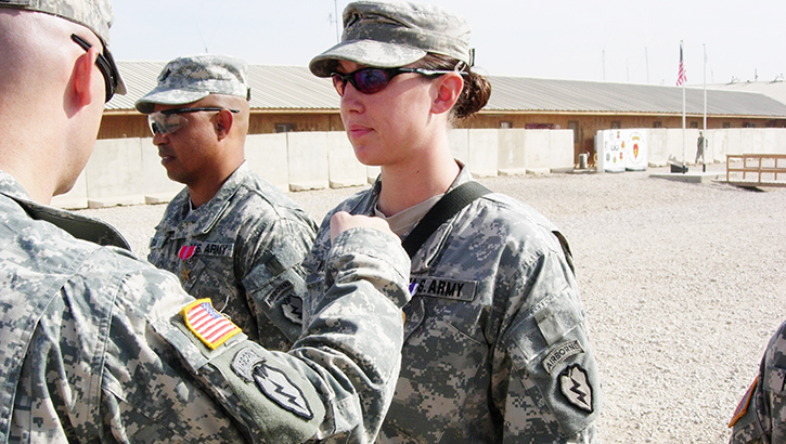 Soldier (center) standing at attention receives an award pinned to their uniform, from a soldier standing directly before her, with a soldier standing at attention to one side. A long building is seen in the background with two flagpoles, one flying the US flag.