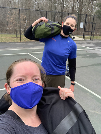Two women, wearing masks, on a tennis court and carrying heavy bundles